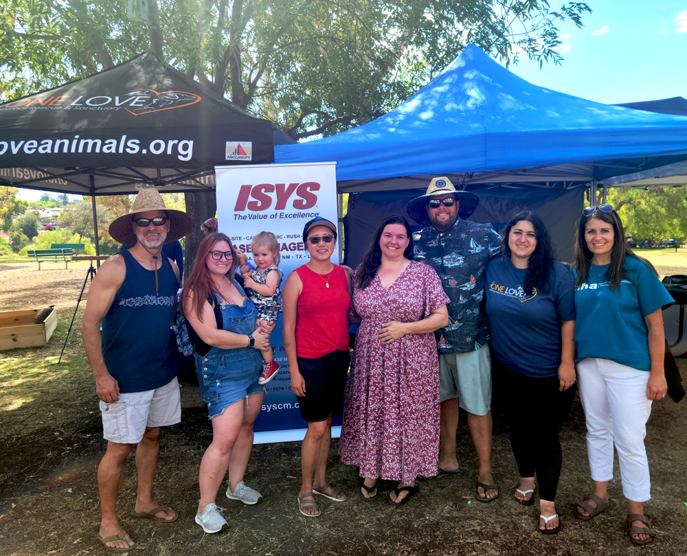 Group of seven adults and one child standing and smiling in front of event tents for One Love Animal Rescue and ISYS at an outdoor park.