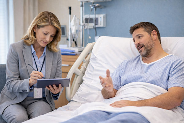 Nurse case manager taking notes on a tablet while speaking with an injured worker in a hospital bed during a workers’ compensation case management visit.