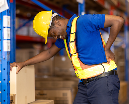 Warehouse worker wearing a yellow hard hat and safety vest holding his lower back in pain, illustrating a workplace back injury that may lead to a workers’ compensation claim.